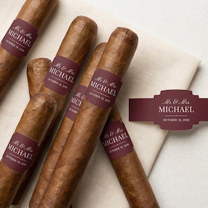 May include: Several brown cigars with burgundy labels that read "Mr & Mrs MICHAEL OCTOBER 18, 2030". A matching label is also present. The cigars are arranged on a white cloth.