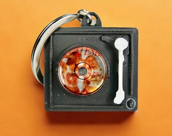 Puede incluir: Un llavero de tocadiscos en miniatura negro con un llavero plateado. El tocadiscos tiene un diseño circular en el centro y un brazo de tono blanco. El fondo es de color naranja liso.