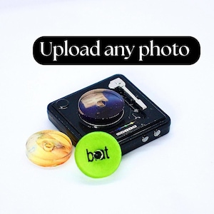 Può includere: Un giradischi in miniatura nero con un braccio argentato e un disco trasparente con un design blu e bianco. Il giradischi è su una superficie bianca con due oggetti rotondi e colorati accanto. Un oggetto è giallo e l'altro è verde con la parola "bot" scritta su di esso.