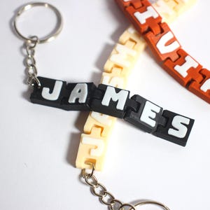 Könnte beinhalten: Schlüsselanhänger mit Namen, darunter "JAMES" in Schwarz und Weiß und "VITA" in Orange und Weiß. Die Schlüsselanhänger bestehen aus ineinandergreifenden Kunststoffteilen und sind an silbernen Schlüsselringen befestigt. Ein weiterer Schlüsselanhänger ist cremefarben.