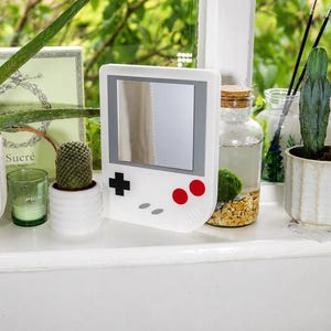 May include: A white handheld gaming console shaped mirror with a square reflective center, a black directional pad, two red circular buttons, and two small gray oval buttons on the front face. The design mimics a classic portable video game system.