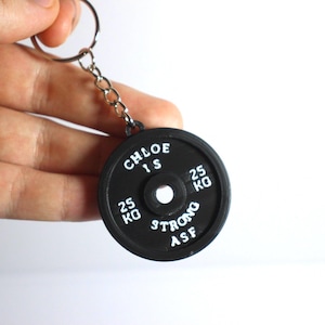 May include: Black weight plate keyring with white lettering. The keyring reads "CHLOE 1S" and "25 KG STRONG ASF". The weight plate has a hole in the centre and is attached to a silver chain. A fitness-themed accessory.