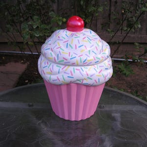 May include: A pink ceramic cupcake cookie jar with white frosting and colorful sprinkles. The jar has a red cherry on top.