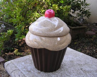 May include: A ceramic cupcake cookie jar with a brown base and a white frosting lid. The lid has a red cherry on top.