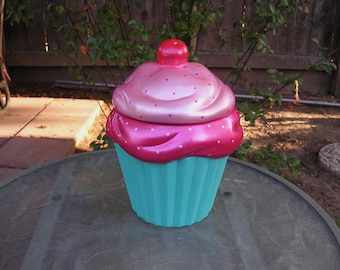 May include: A pink and turquoise ceramic cupcake cookie jar with white polka dots. The lid is shaped like a cupcake with a pink frosting swirl and a red cherry on top.