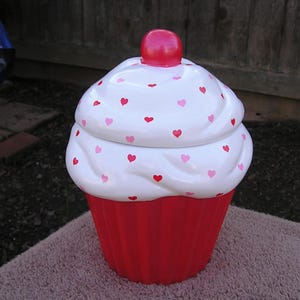 May include: A white ceramic cupcake cookie jar with a red and white frosting design and pink and red hearts. The lid is shaped like a cupcake with a red cherry on top.