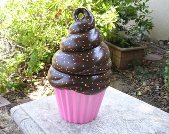 May include: A whimsical cupcake-shaped container with a pink base and chocolate frosting top. The brown frosting is decorated with colorful sprinkles. The container is sitting on a stone surface, with greenery in the background.