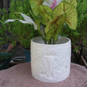 May include: White ceramic planter with an embossed design of elephants and foliage. The planter is filled with green and white leafy plants.