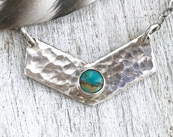 May include: A chevron-shaped silver pendant featuring a hammered texture and a single round turquoise stone set in the center. The pendant is attached to a thin silver chain, creating a necklace that displays a metallic sheen and blue-green color.