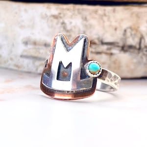 May include: A silver ring with a copper band featuring a cutout letter "M" and a turquoise stone.