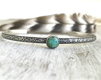May include: A silver cuff bracelet featuring a central round turquoise stone with natural brown matrix patterns. The band has a dark oxidised finish and is decorated with a repeating embossed floral and geometric pattern along the entire length.