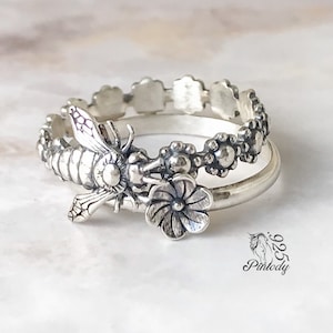 May include: A silver ring set with a bee and flower design. The ring is made of two bands, one with a floral pattern and the other with a bee design.