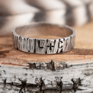 May include: A silver ring with a textured, bark-like finish featuring the stamped letters L and C separated by a small plus sign. The band has a rugged, uneven surface that mimics natural wood grain, providing a distinct tactile appearance.