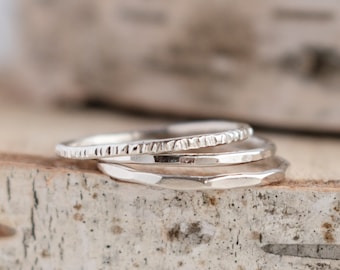 May include: A set of three stacked silver rings featuring distinct textures. The top ring has a fine vertical etched pattern, the middle ring has a smooth polished surface, and the bottom ring displays a faceted, hammered finish with reflective edges.