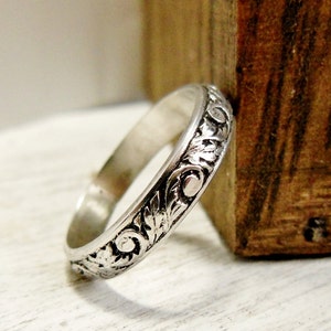 Pode incluir: Um anel de banda de prata com um design floral detalhado. O anel est&aacute; sobre uma superf&iacute;cie de madeira.