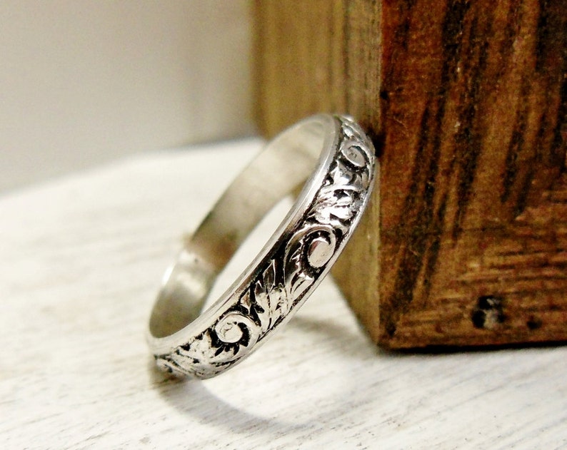 Pode incluir: Um anel de banda de prata com um design floral detalhado. O anel est&aacute; sobre uma superf&iacute;cie de madeira.