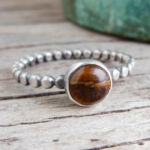 May include: A silver ring with a round, polished tiger's eye stone. The band is made of small, evenly spaced silver beads. The ring is set against a weathered wooden surface.
