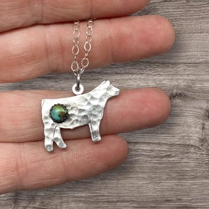 May include: A silver cow pendant necklace with a hammered texture. The cow has a turquoise stone accent. The necklace chain is a delicate silver. The pendant is held between fingers.