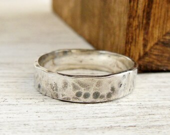 May include: A silver ring with a hammered, textured surface. The band has a slightly uneven, organic edge and a metallic finish that reflects light across its dimpled exterior, showcasing a handcrafted appearance with visible indentations.