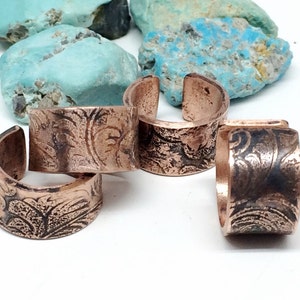 May include: Four copper rings with an etched floral design. The rings are stacked on top of each other.