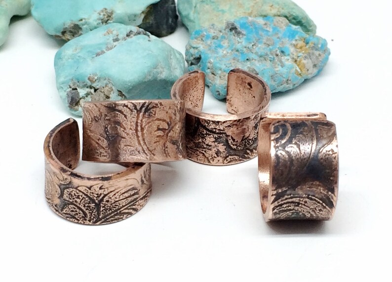 May include: Four copper rings with an etched floral design. The rings are stacked on top of each other.