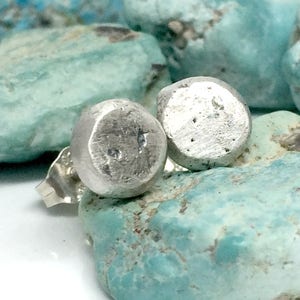 May include: Two small, silver stud earrings with a textured, rounded design. The earrings are presented against a backdrop of light blue, rough-textured stones. The earrings have a simple, minimalist style.