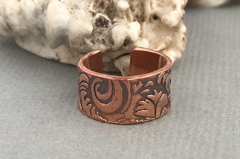 May include: A copper ring with a wide band and an intricate floral design. The ring is open on one side and has a smooth, polished finish.
