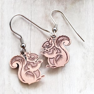 May include: A pair of copper earrings featuring a detailed engraved design of a squirrel. The earrings have silver hooks.