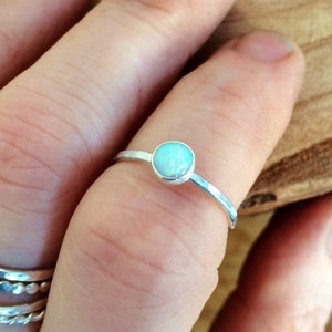 May include: A thin silver ring featuring a small, round, iridescent opal stone set in a bezel. The band has a smooth, polished finish. Below this ring, two additional silver bands are visible, one with a textured surface and one with a twisted design.