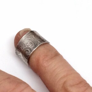 May include: A silver ring with a textured, swirling design. The ring is worn on a finger.