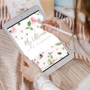 May include: A white tablet displaying a planner design with pink roses, green leaves, and the text "My Ultimate Life Planner." A white stylus is held over the screen. The tablet is held by a person.