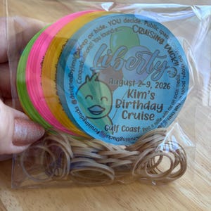 May include: A stack of circular neon-colored tags in a clear plastic bag with a pile of tan rubber bands. The top tag is light blue with a cartoon duck, balloons, and text reading Liberty, August 2-9, 2026, Kim's Birthday Cruise, Gulf Coast.