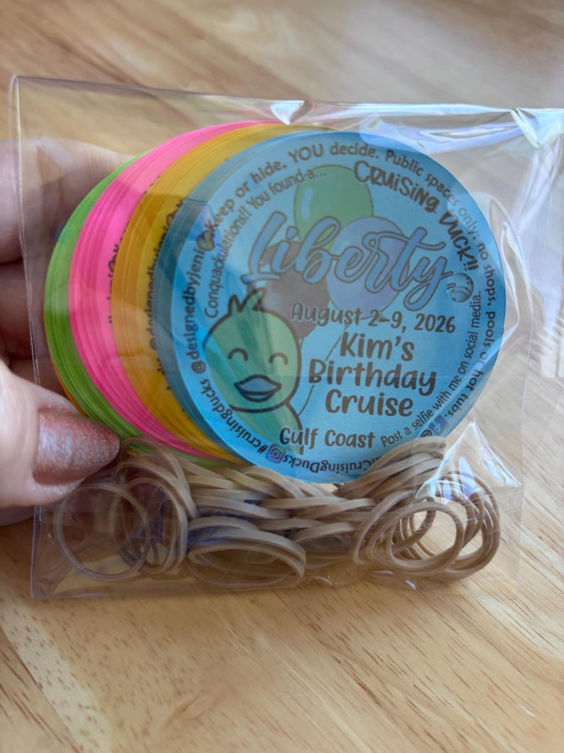 May include: A stack of circular neon-colored tags in a clear plastic bag with a pile of tan rubber bands. The top tag is light blue with a cartoon duck, balloons, and text reading Liberty, August 2-9, 2026, Kim's Birthday Cruise, Gulf Coast.