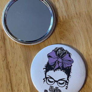May include: A white compact mirror with a purple and black illustration of a woman with a bun and glasses. The text "Hello Beautiful" is printed below the illustration.