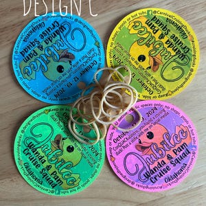 May include: Four circular paper tags in blue, yellow, green, and pink, each featuring a cartoon duck, the text Jubilee, Wanda & Pam Cruise Squad, Oklahoma, and October 12-19, 2024, with a small hole and a cluster of tan rubber bands in the center.