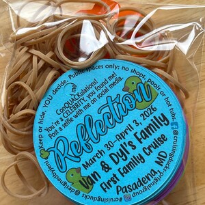 May include: A clear plastic bag filled with tan rubber bands and a round blue tag. The tag features green duck illustrations and text: Reflection, March 30-April 3, 2026, Van & Dyl's Family First Family Cruise, Pasadena, MD, and social media handles.