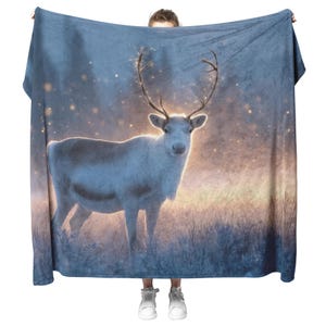 May include: A large, rectangular blanket featuring a majestic white reindeer with large antlers. The reindeer stands in a field with a soft, glowing background of blue and gold. The blanket is held up, showcasing the full design.