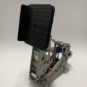 Könnte beinhalten: Ein silbernes Rennpedal aus Metall mit einer schwarzen Fußstütze und einem Federmechanismus. Das Pedal ist für die Verwendung mit einem Rennsimulator konzipiert.