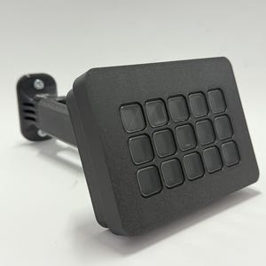 Könnte beinhalten: Eine schwarze Tastatur aus Kunststoff mit 16 quadratischen Tasten. Die Tastatur ist an einem schwarzen Kunststoffarm befestigt.