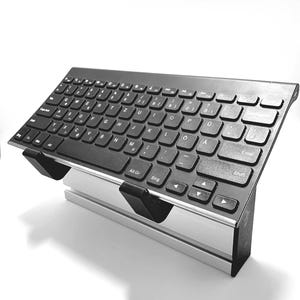 Op de afbeelding: Zwart draadloos toetsenbord met een volledige QWERTY-indeling, op een zilverkleurige standaard. Het toetsenbord heeft zwarte toetsen met witte letters. De standaard biedt een schuine typpositie.