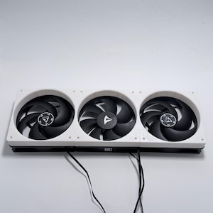 Könnte beinhalten: Eine weiße, rechteckige Kühlventilatoreinheit mit drei schwarzen Lüftern. Jeder Lüfter hat gebogene Flügel und ein zentrales Logo. Die Einheit ist für die Computerkühlung konzipiert.