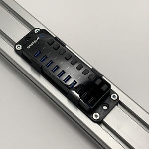 Op de afbeelding: Een zwarte USB-hub met meerdere poorten en de merknaam "SABRENT" erop gedrukt. De hub is gemonteerd op een zilveren metalen rail met schroeven. De hub heeft blauwe USB-poorten.