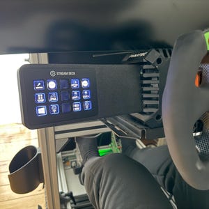 Può includere: Un pannello di controllo Stream Deck nero con pulsanti illuminati, montato vicino a un volante da corsa. Il volante ha un'impugnatura nera e un design in fibra di carbonio. L'immagine mostra una configurazione di gioco.