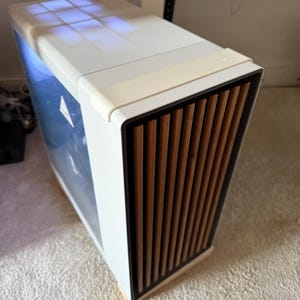 Può includere: Un case per computer bianco con un pannello frontale in legno e una parte superiore a rete. Il case ha un pannello laterale trasparente. Il pannello frontale presenta doghe di legno verticali. Il design è moderno ed elegante.