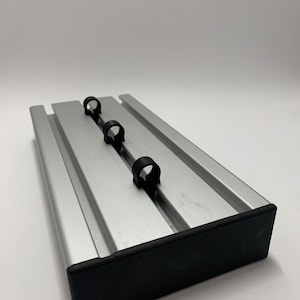 Op de afbeelding: Een zilverkleurige metalen basis met drie zwarte plastic clips. De basis heeft een rechthoekige vorm met groeven en een zwarte rubberen rand. De clips zijn gelijkmatig verdeeld.