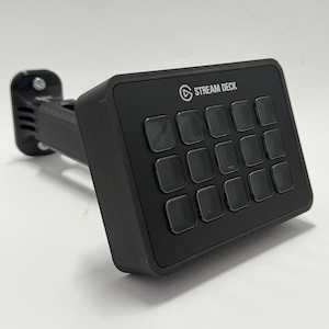 Könnte beinhalten: Ein schwarzes Stream Deck mit 15 quadratischen Tasten und dem Stream Deck-Logo. Das Gerät ist an einem schwarzen, verstellbaren Arm mit einer Halterung befestigt.