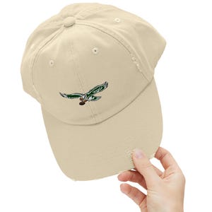 May include: A beige distressed baseball cap with a green and white embroidered eagle logo.