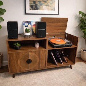 May include: A mid-century modern style record player console in a warm wood tone. The console features a turntable with an orange record, two black speakers, and storage for vinyl records. Decorative plants and a floor lamp are also visible.