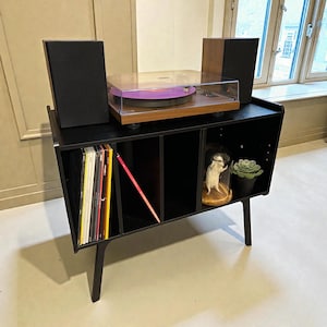 May include: A black record player stand with a turntable, speakers, and storage for vinyl records. The turntable has a clear cover and a purple record. A small glass dome with a figurine and a potted succulent are on the shelf.