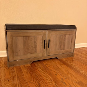 May include: A wooden shoe bench with two doors and a padded top. The bench is made of light brown wood with a textured grain. The doors have black handles. The padded top is a dark gray color.